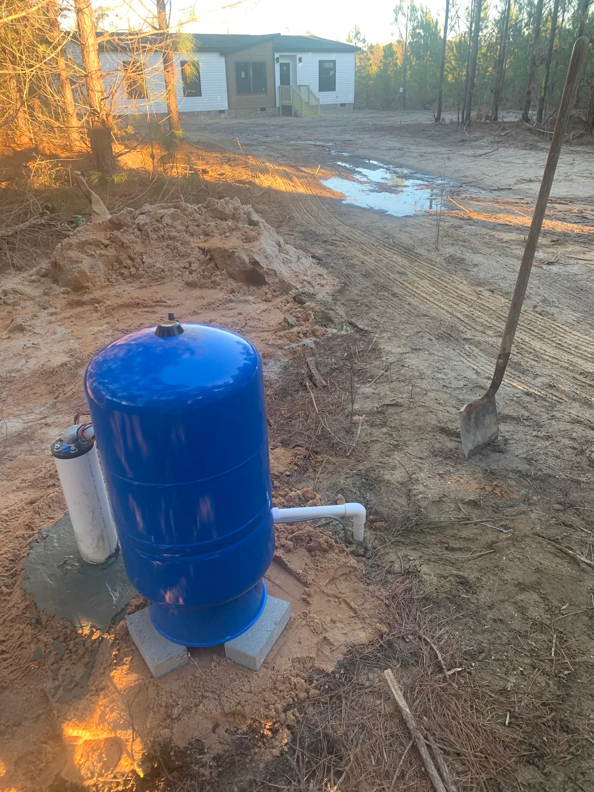 Well pressure tank and piping with a manufactured home in the background.